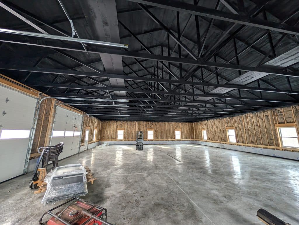 PXL_20251215_180400528 Large garage interior with exposed insulation and painted black ceiling rafters, ready for services.