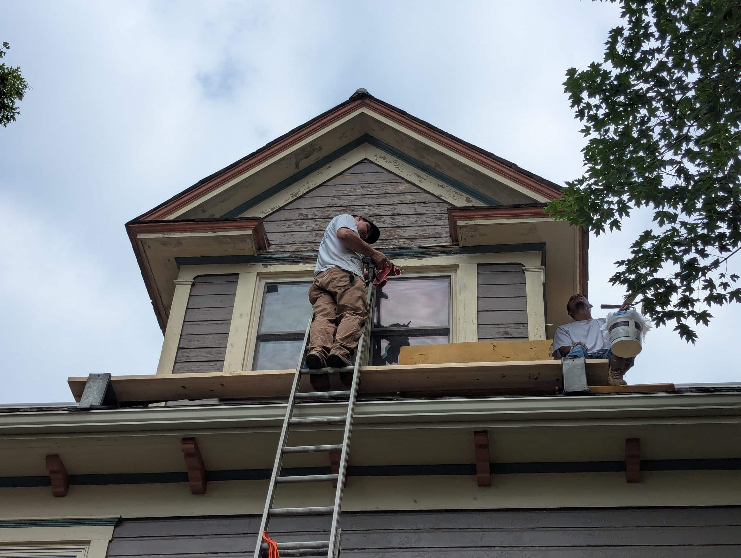 Painters working on the exterior of a brown and beige house, on scaffolding.