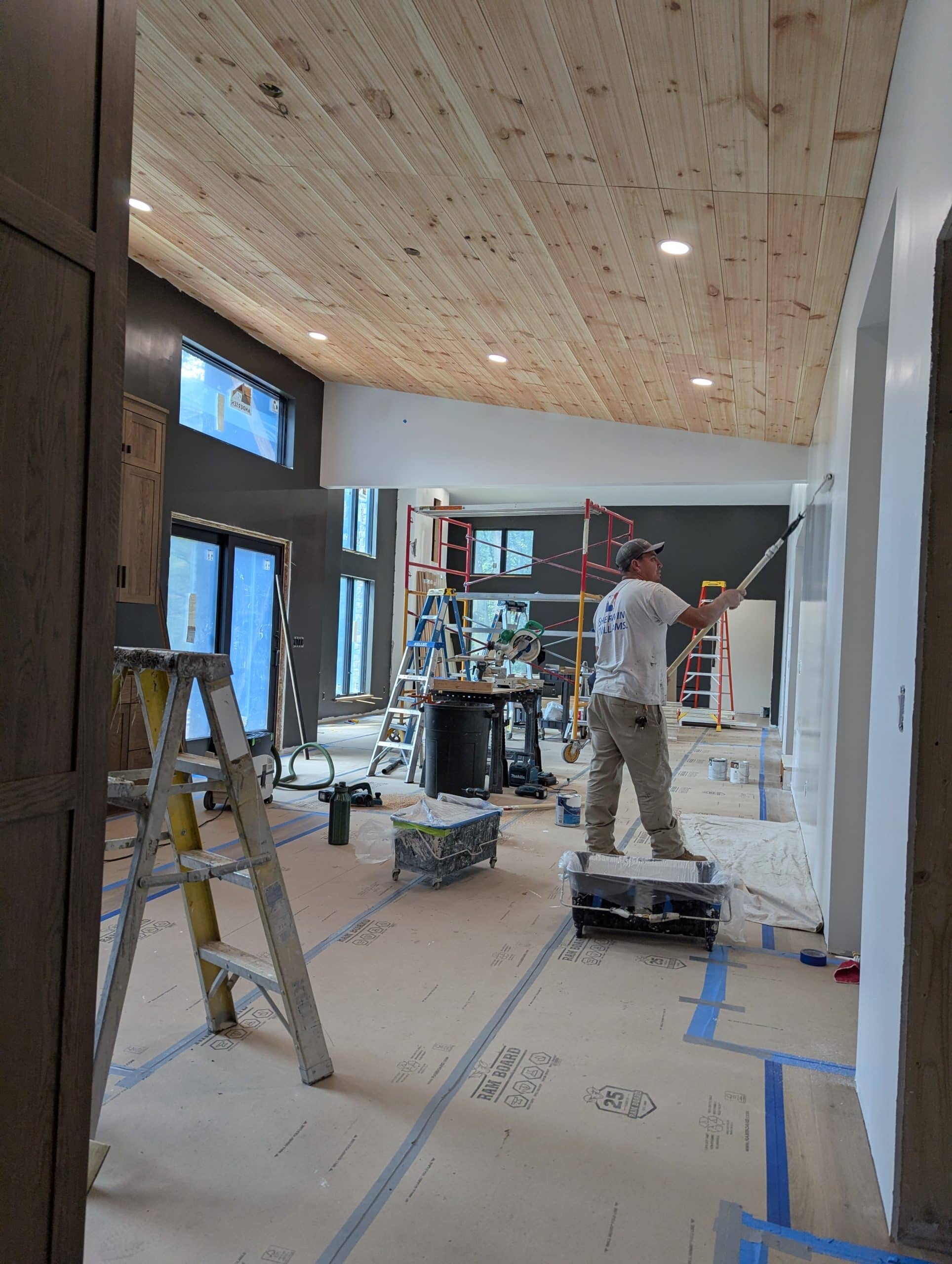 Painter using roller on interior wall, ladders and equipment visible.