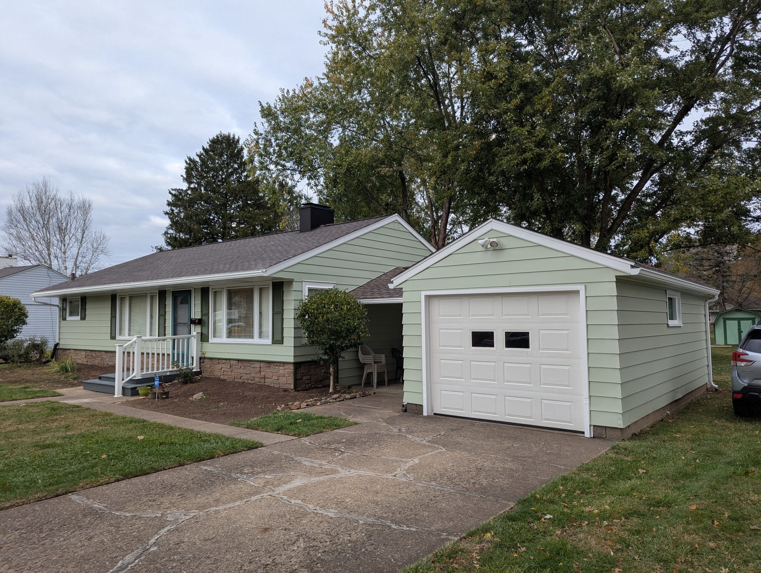 Light green ranch style house with stone foundation and detached garage on a cloudy day.